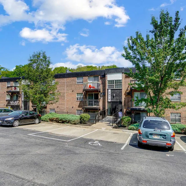 View of a brick apartment complex surrounded by greenery and a parking lot. The sky is partly cloudy.
