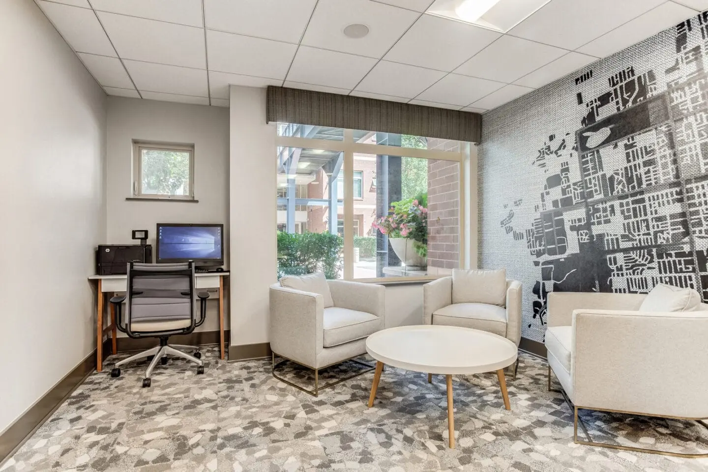 Modern Lounge Area A modern lounge area featuring two armchairs and a round coffee table, a desk with a computer in the corner, and a large window with a view of outdoor greenery. The wall behind has a graphic design, and the flooring consists of patterned tiles.