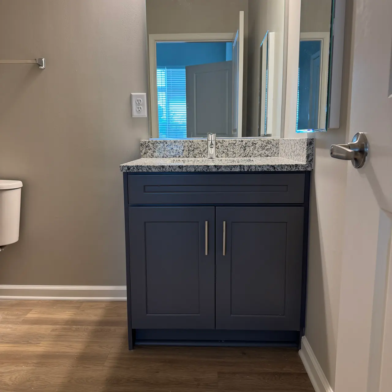 A modern bathroom featuring a dark blue vanity with a granite countertop and a large mirror above. The wall behind is painted in a neutral tone. Light wooden flooring is visible, and a white toilet is located to the left.
