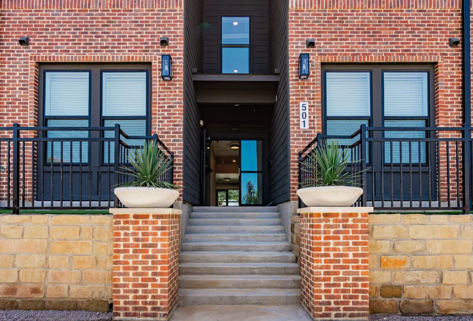 The Gleason A modern brick building entryway featuring two large windows, a double-door entrance, and two planters with plants on either side of the stairs.