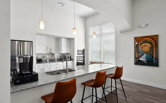 A modern kitchen with a breakfast bar featuring four brown leather stools. The kitchen has white cabinetry, stainless steel appliances, and a granite countertop. Large windows provide natural light, and artwork is visible on the wall.