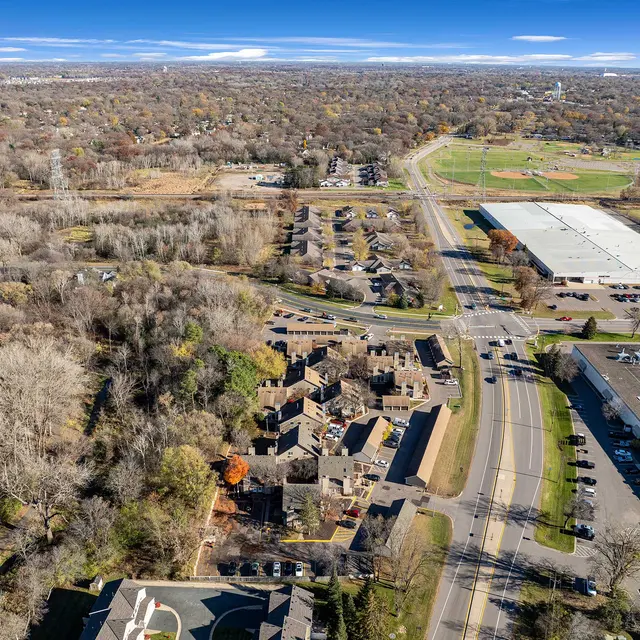Aerial view of a suburban area featuring residential buildings, a wide road, and patches of forest. In the background, there is a commercial building and an open green field.