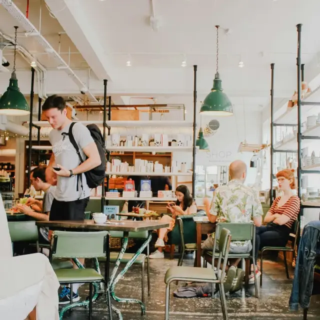 Social Scene in a Cozy Cafe A light-filled cafe interior featuring various people seated and standing around tables, some looking at their phones and others engaged in conversation. The decor includes green pendant lights and bookshelves filled with products.