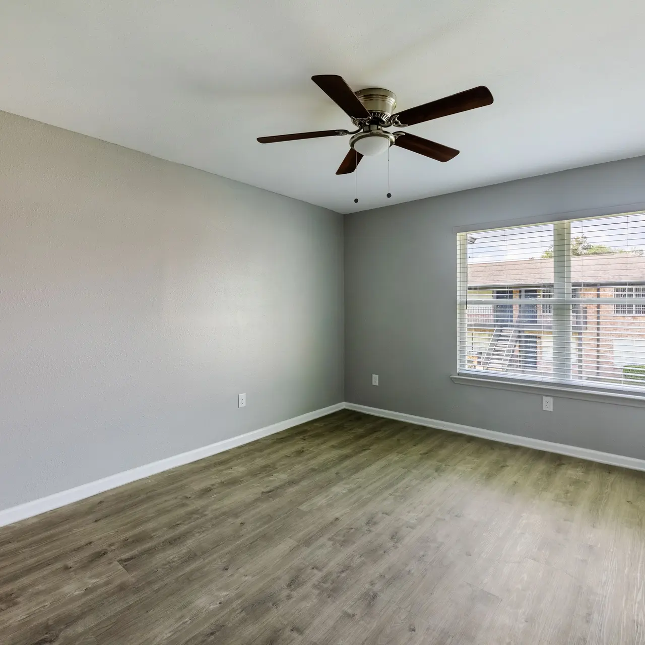 An empty room with a light gray wall and a ceiling fan. A large window with blinds allows natural light to enter, casting a bright appearance on the wooden floor.