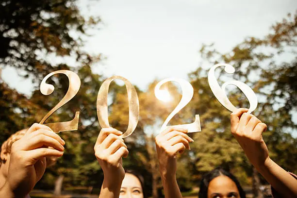 A group of hands holding up golden numbers that spell out '2026' against a bright, outdoor background with trees.