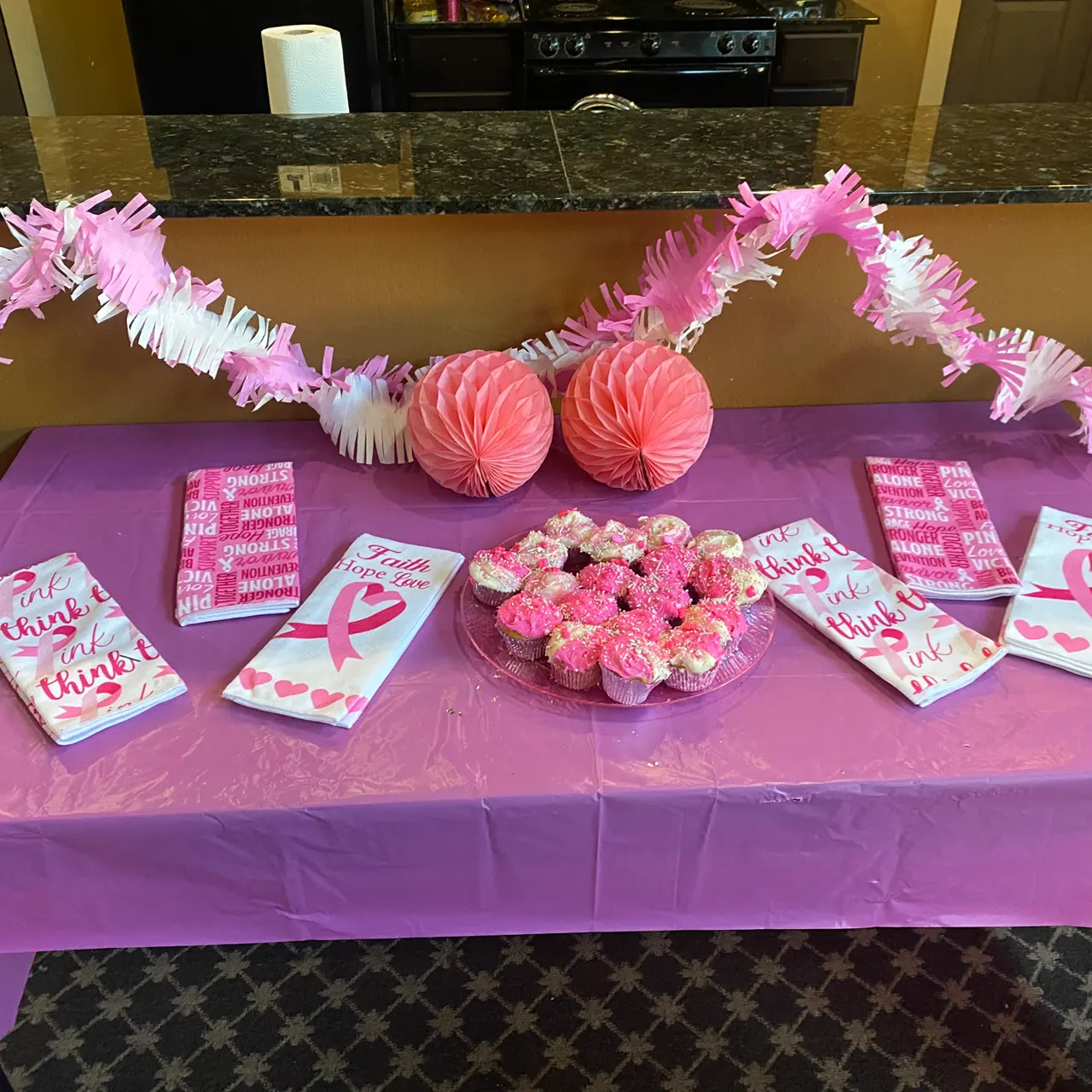 A table decorated with pink tablecloth and items for a celebration, featuring pink garlands, cups, and a plate of pink frosted cupcakes.