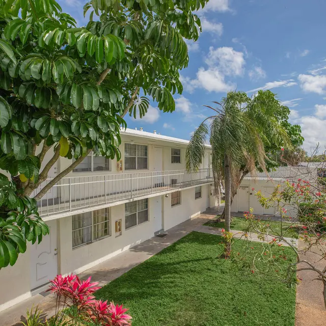View of an apartment complex surrounded by green trees and plants, with a sunny sky in the background.
