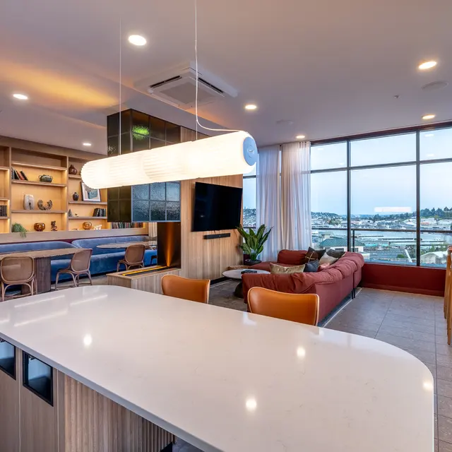 A contemporary living space featuring a white island countertop, seating area with a red couch, and large windows offering a view. Decor includes shelves with books and decorative items, and stylish lighting.