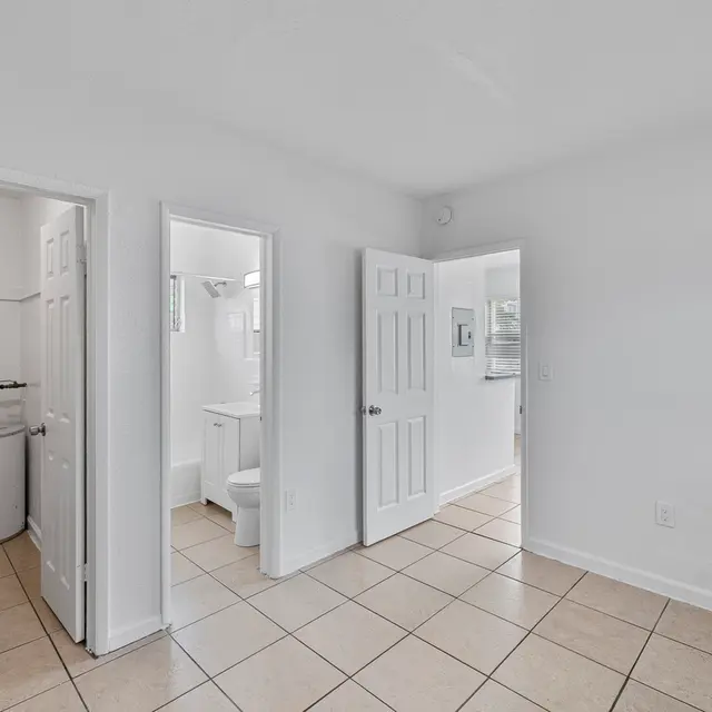A clean and bright interior of a bathroom area with white walls and tiled flooring. There are three doors leading to different spaces, including one that appears to lead to a laundry area with a water heater visible. The second door leads to a bathroom with a sink and toilet accessible, and the third door opens to another room with a window.