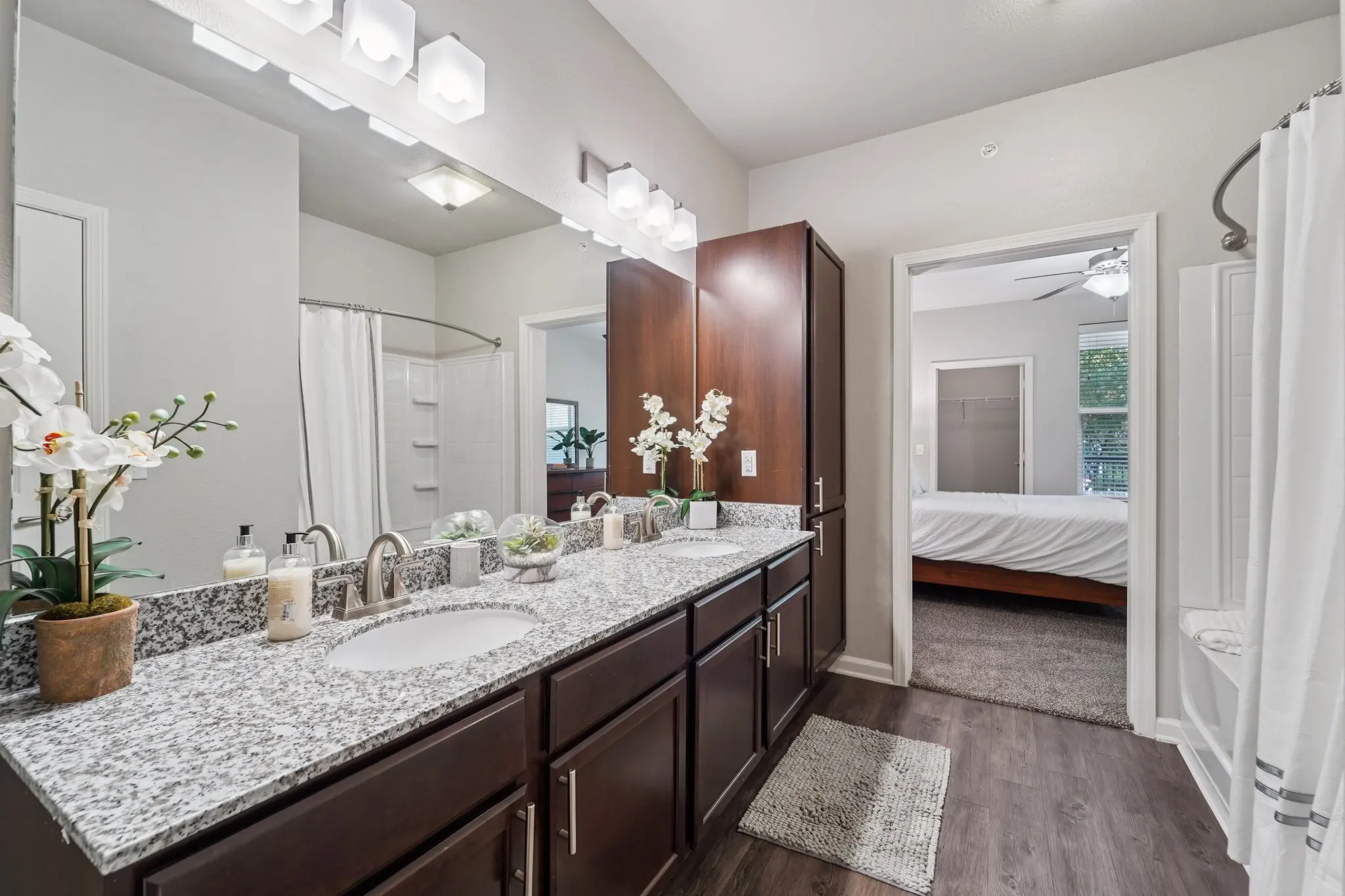 A modern bathroom featuring a double vanity with a stone countertop, white cabinetry, and decorative plants. A shower area is visible on the left with a curtain. A doorway leads to a bedroom in the background, showing a bed with neutral bedding. The flooring is a dark wood finish and the walls are painted in a light color.
