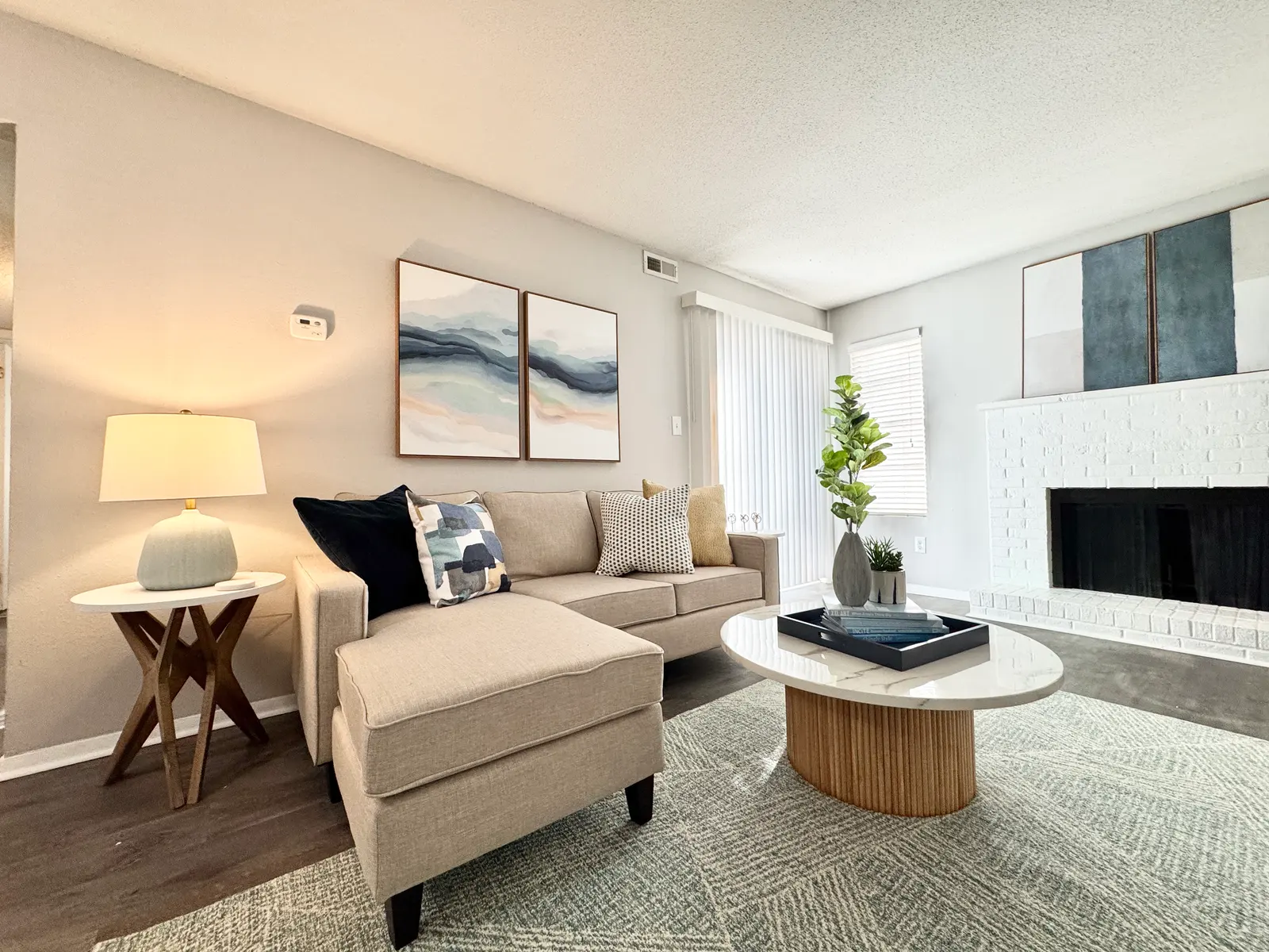 A stylish living room featuring a beige sectional sofa, a light-colored coffee table with a round wooden base, and an elegant lamp. The walls are painted in a light shade, adorned with abstract art, and there are blinds on the windows with natural light streaming in.