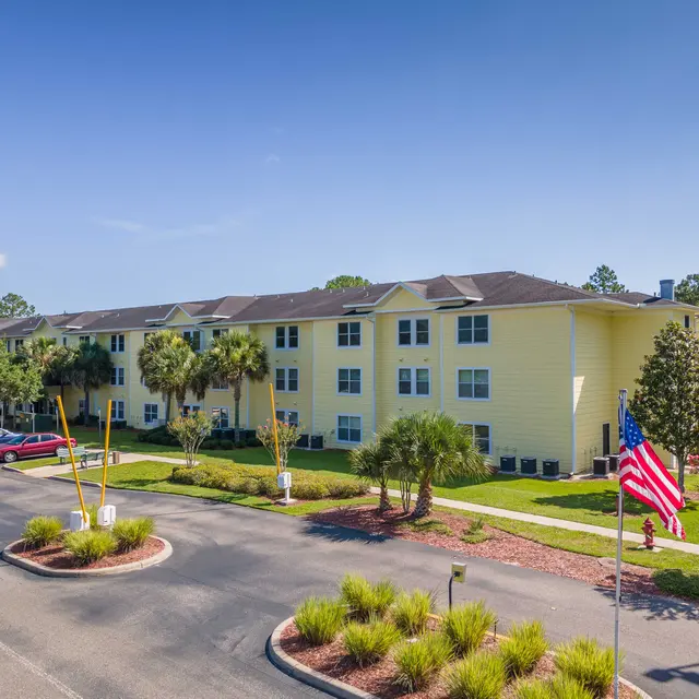 A bright yellow three-story apartment building surrounded by greenery and palm trees, with a parking lot in the foreground. An American flag is flying near the entrance of the building.
