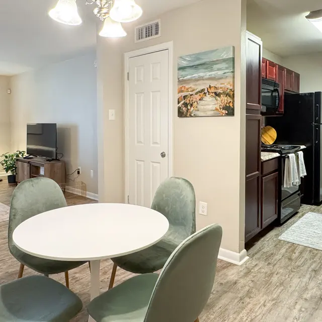 A well-lit living space showing a dining area with a round white table and four green chairs, a kitchen with dark wood cabinets, and a comfortable living area with a television and a sofa in the background.