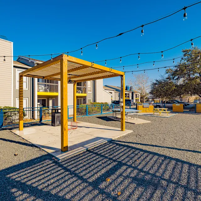 A bright outdoor space featuring a wooden structure with a pergola, surrounded by string lights and gravel. There are benches and grassy areas in the background, accompanied by residential buildings.