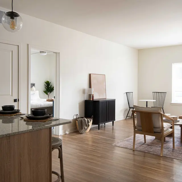 A modern, minimalistic living space featuring a kitchenette, dining area, and lounge area. Natural light floods the room through a large window, highlighting the wooden floors and contemporary furniture.