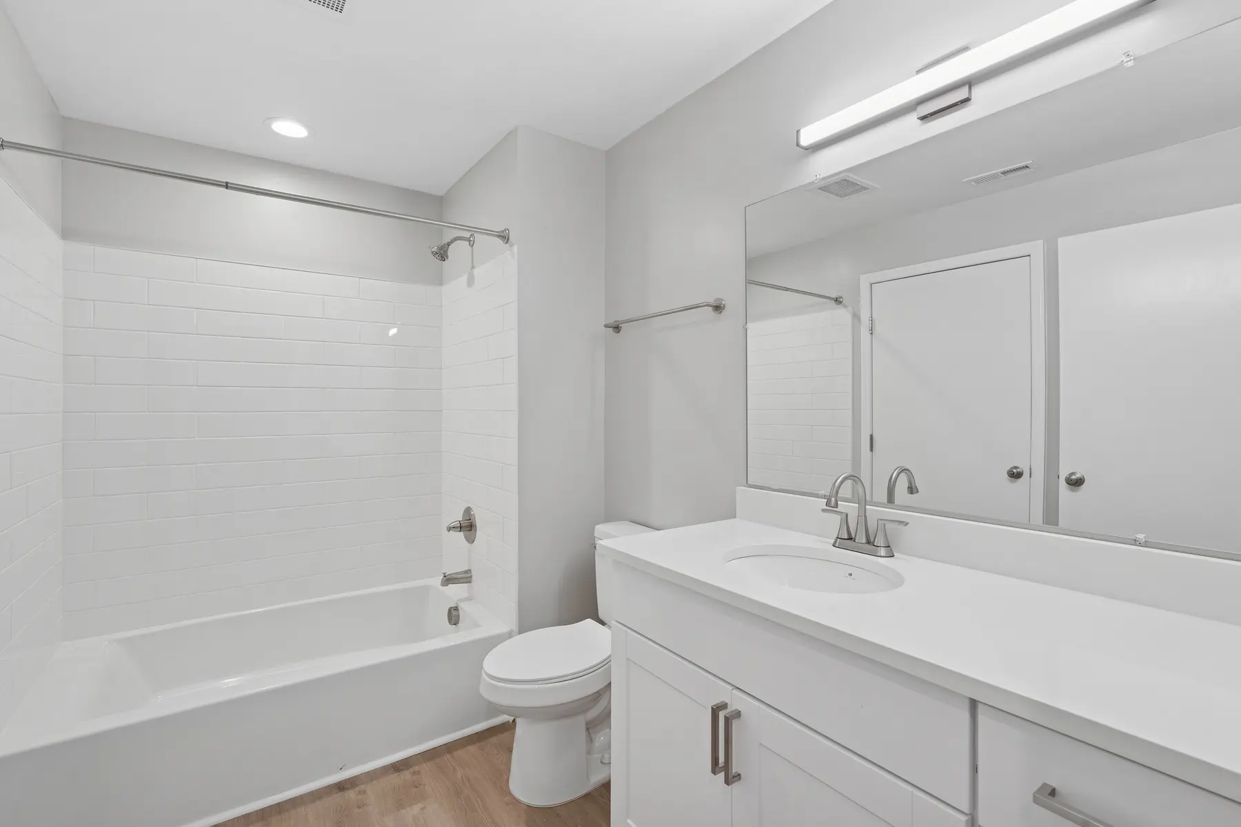 A modern bathroom featuring a large bathtub with a shower, a white vanity with a sink, and a mirror above it. The walls are painted in a light color and there is wood flooring.