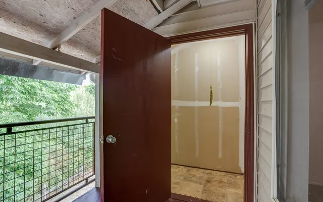 An open maroon door leading to an unfinished space, with partially painted walls and a view of greenery outside through a balcony railing.