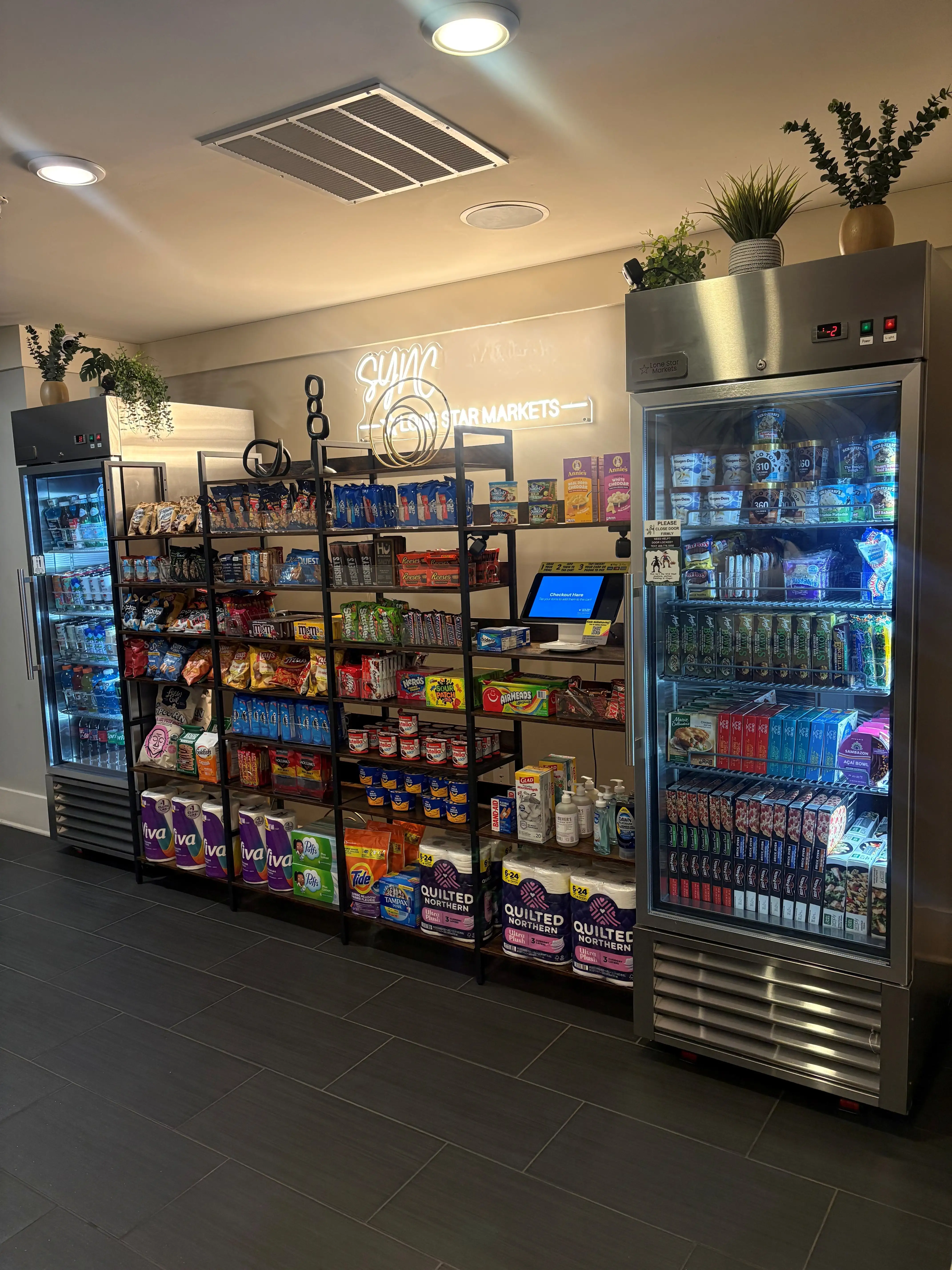 A modern community market area with shelves stocked full of snacks, drinks, and essential items, including two glass refrigerated units.