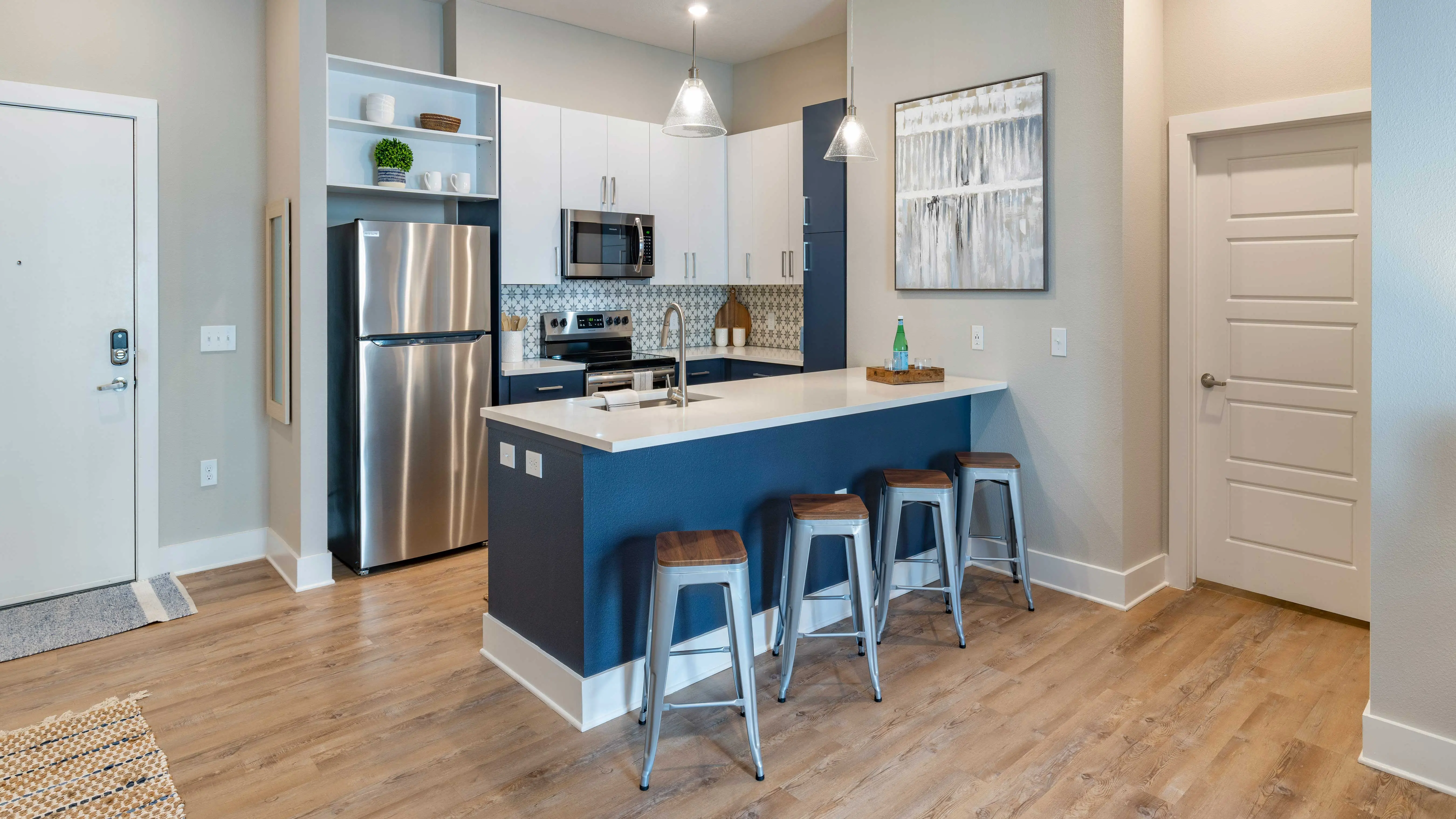 Modern kitchen featuring a stainless steel refrigerator, microwave, and oven. The island has seating for three with wooden stools. Blue and white cabinetry, and wood flooring are present.