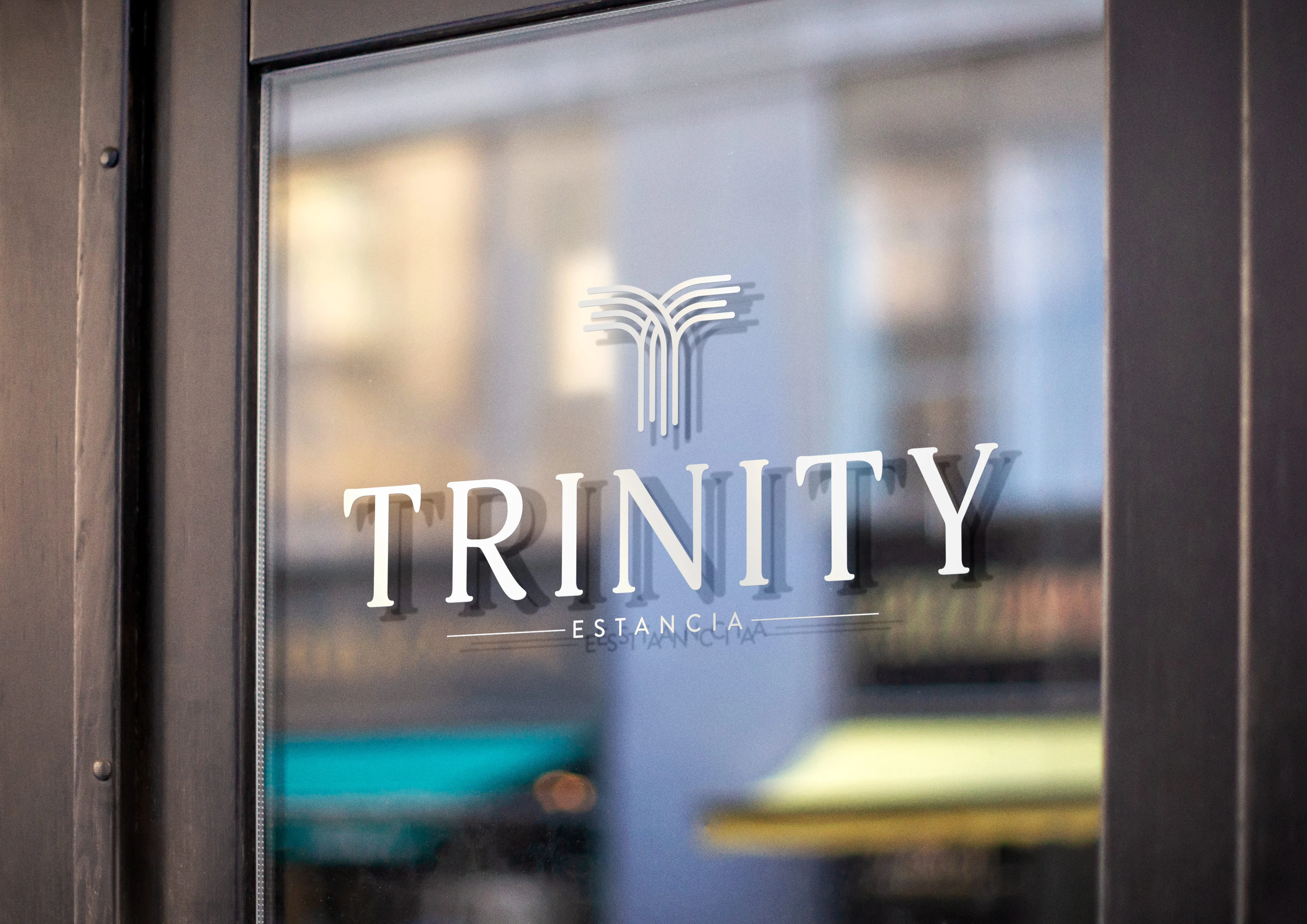 Trinity Estancia Logo A close-up view of a glass window featuring the logo of 'Trinity Estancia'. The logo is elegantly designed with the word 'TRINITY' in a bold font, while 'ESTANCIA' is in a smaller, subtler font below it. The background is softly blurred, highlighting the logo.