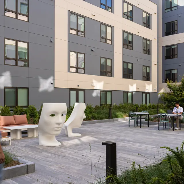 A contemporary courtyard featuring unique art installations of abstract heads, surrounded by greenery and seating areas. A woman walks past the art while a man sits at a table in the background.