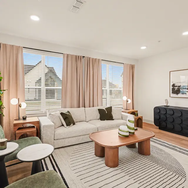A modern living room featuring a light-colored sofa, round coffee table, green accent chairs, a plant, and a decorative sideboard. Large windows allow natural light to fill the space, and curtains frame the windows.