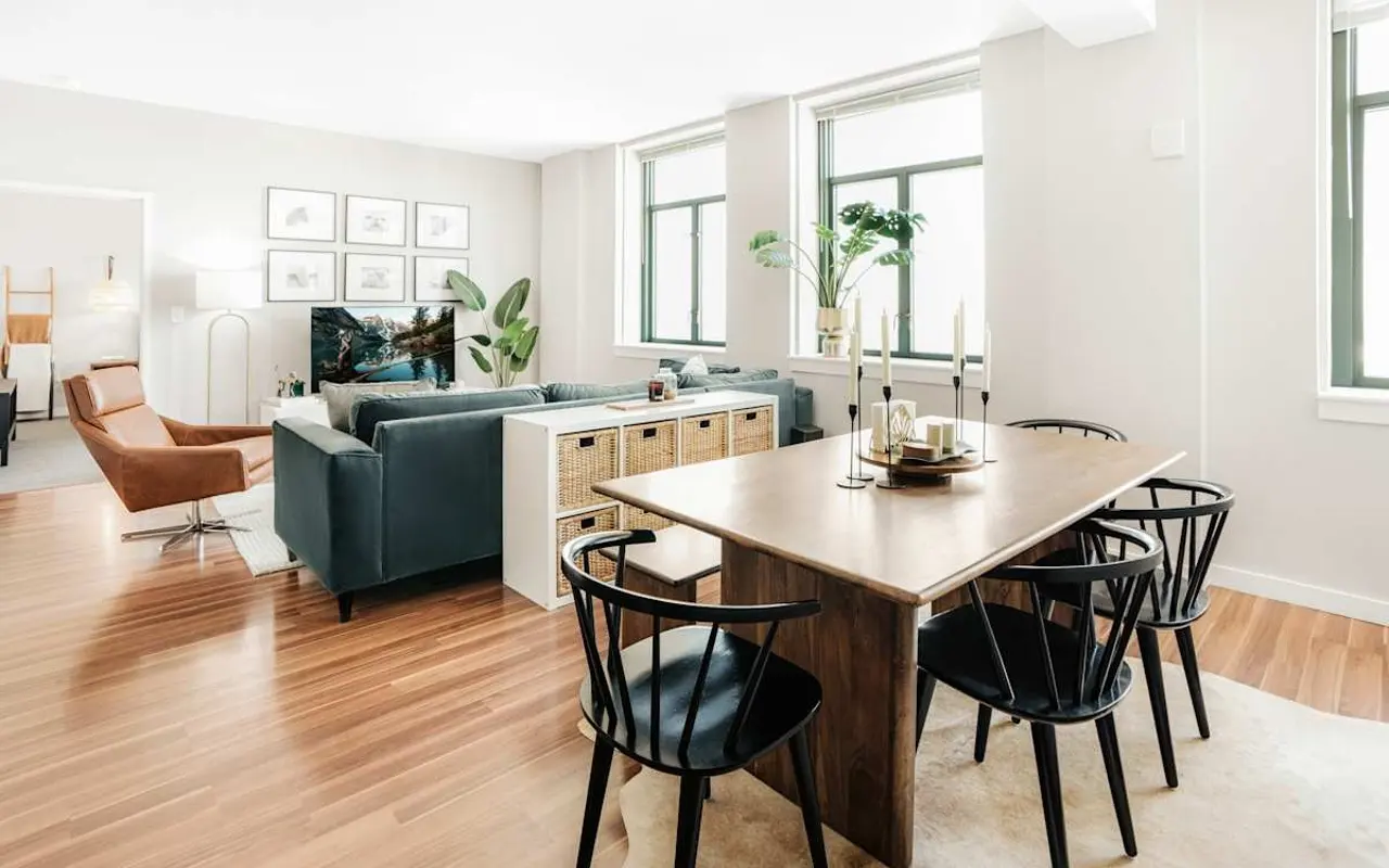 A modern living and dining area featuring a wooden dining table surrounded by black chairs, a green sofa, and a cozy brown armchair. Large windows allow natural light to fill the space, complemented by decorative plants and wall art.