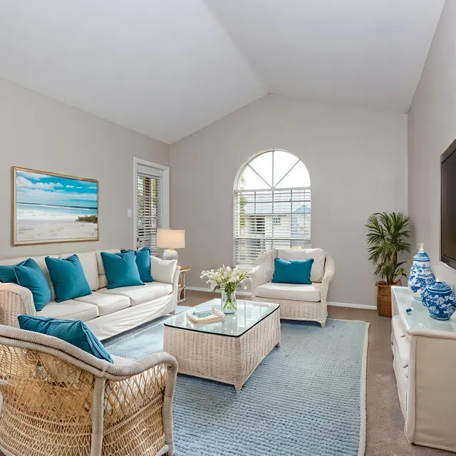 A bright and airy living room featuring a comfortable sofa with turquoise cushions, a glass coffee table, and a TV mounted on the wall. There are two lamps on either side of the sofa, and a decorative plant in the corner. A painting with a beach scene hangs on the wall, next to a large window allowing natural light to fill the space.