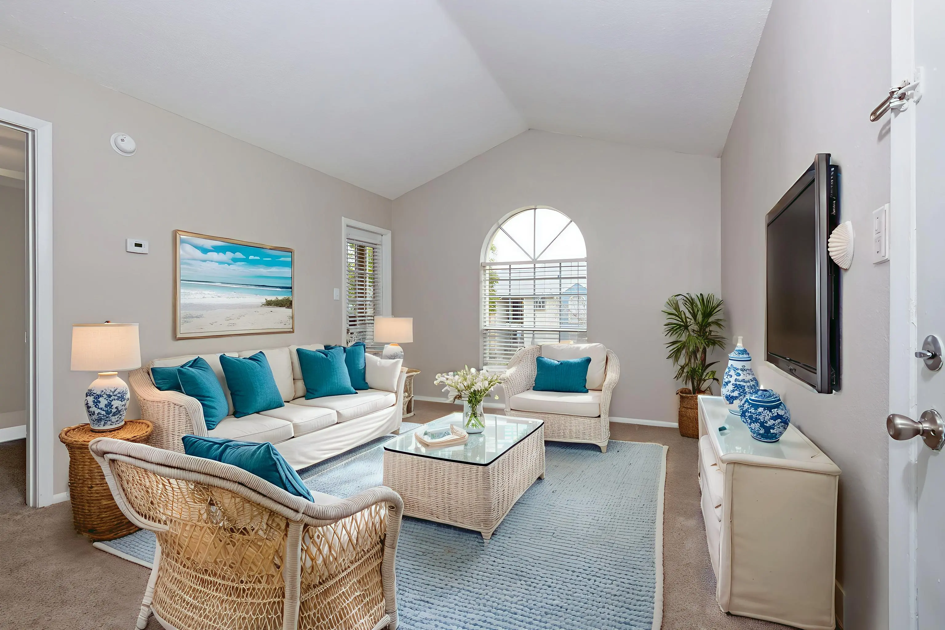 A bright and airy living room featuring a comfortable sofa with turquoise cushions, a glass coffee table, and a TV mounted on the wall. There are two lamps on either side of the sofa, and a decorative plant in the corner. A painting with a beach scene hangs on the wall, next to a large window allowing natural light to fill the space.