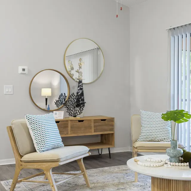 A modern living room featuring two stylish chairs with decorative cushions, a round wooden coffee table, and a console table adorned with a mirror and decorative items. The room has light grey walls and large windows with vertical blinds, creating a bright and airy atmosphere.