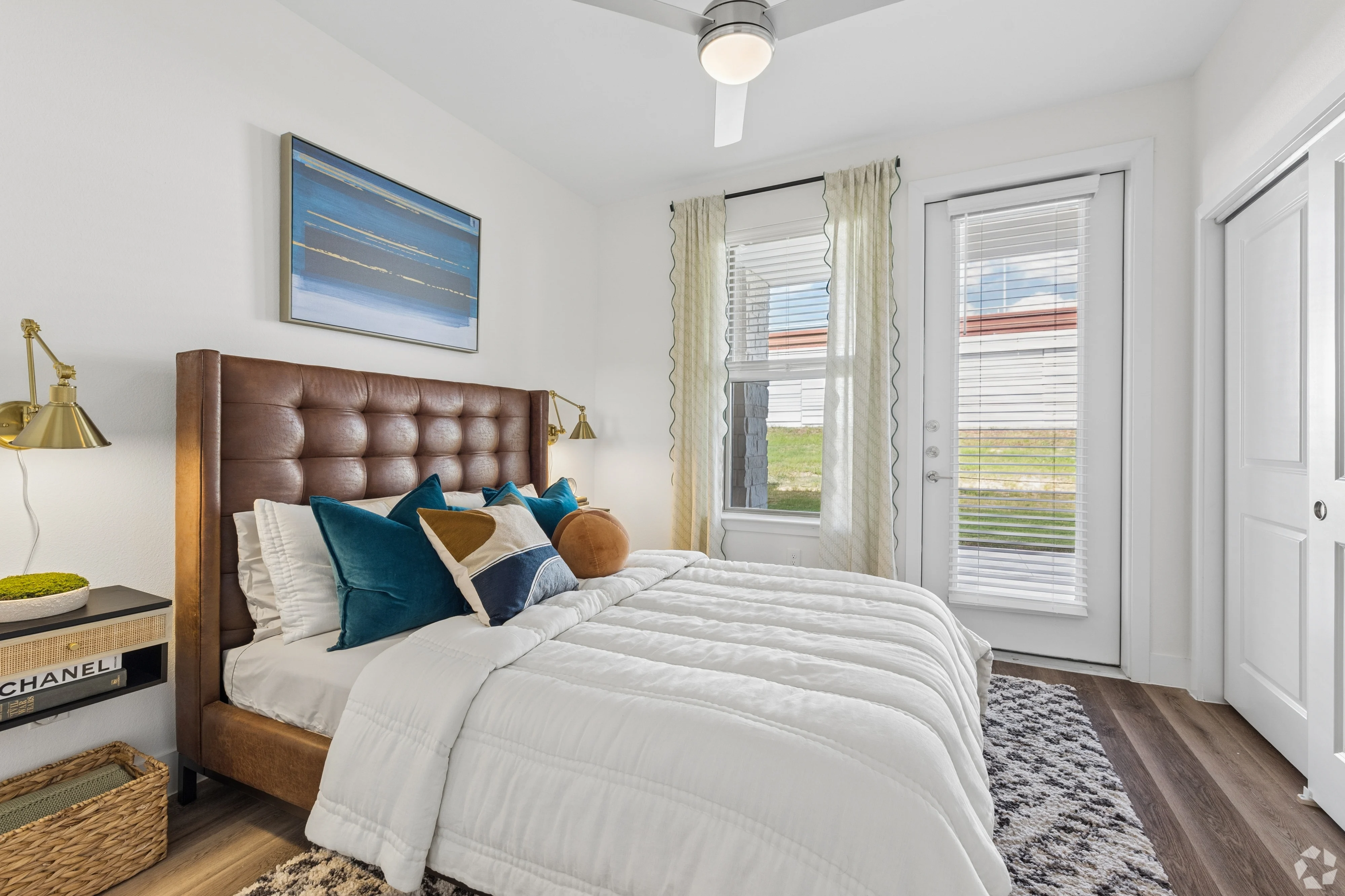 A modern bedroom featuring a brown tufted headboard bed with white linens and decorative pillows, two windows with white curtains, and a stylish nightstand with a lamp and decorative items.