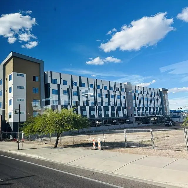 A modern, multistory building with a distinct architectural design, featuring a mix of blue, gray, and white panels. There are shrubs and a small tree in front, and a clear blue sky with scattered clouds above.