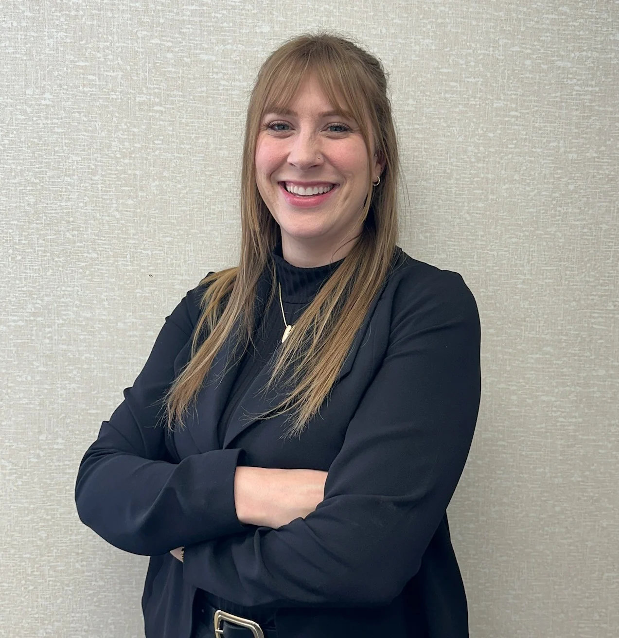 A woman standing with her arms crossed, smiling, wearing a black blazer and a black turtleneck top, against a light gray background.