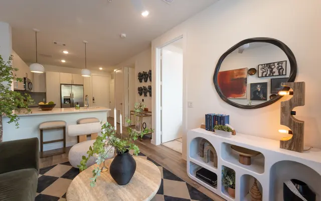 A modern apartment living room featuring a cozy seating area with a round wooden coffee table, green chairs, and a decorative vase with plants. The background shows a stylish kitchen with stainless steel appliances and a wall decorated with framed art. The room is bright with neutral colors and natural light.
