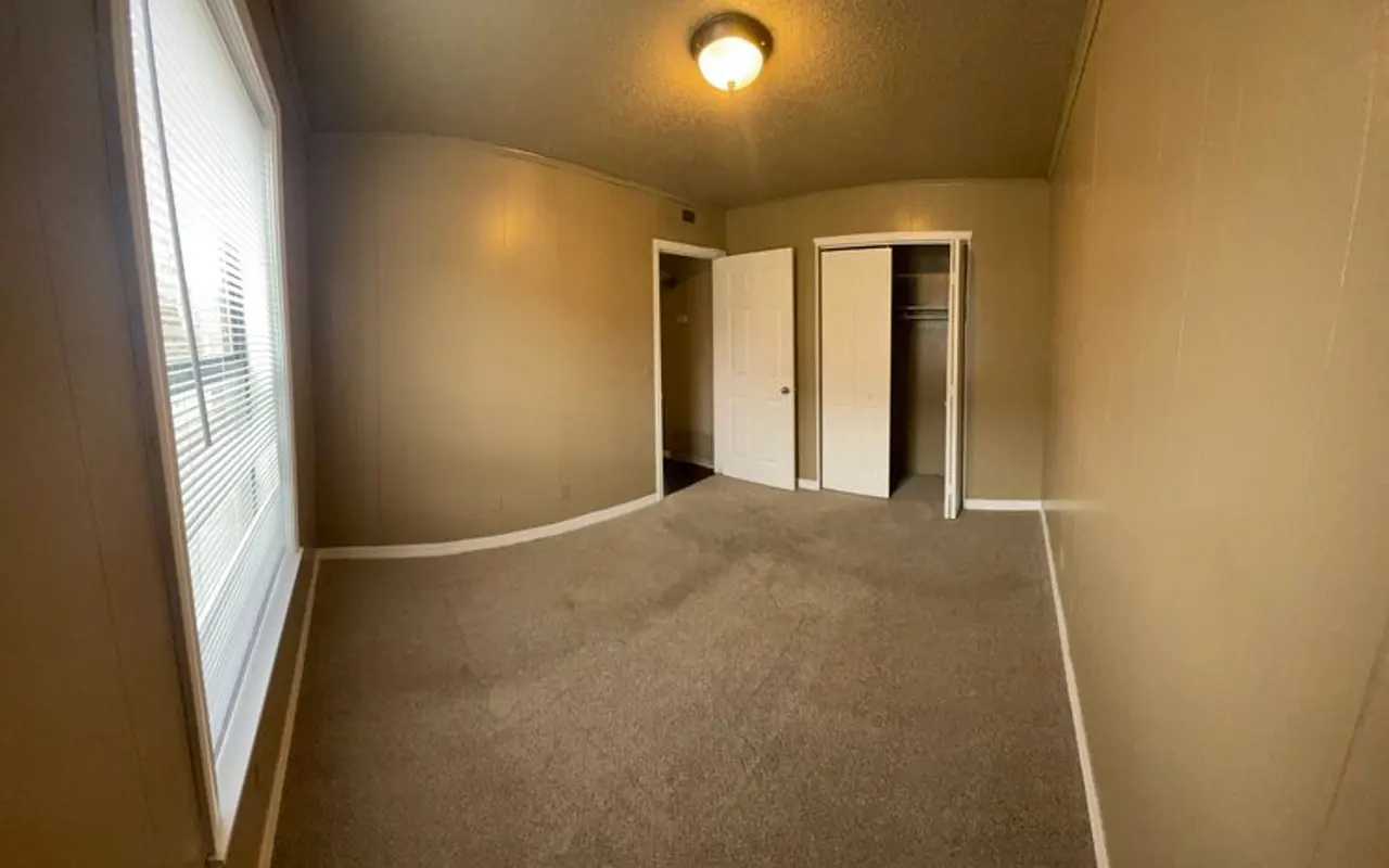 Empty Room Interior An empty room with beige walls, a carpeted floor, and a window with blinds letting in natural light. There are two doors leading to a closet and another space.