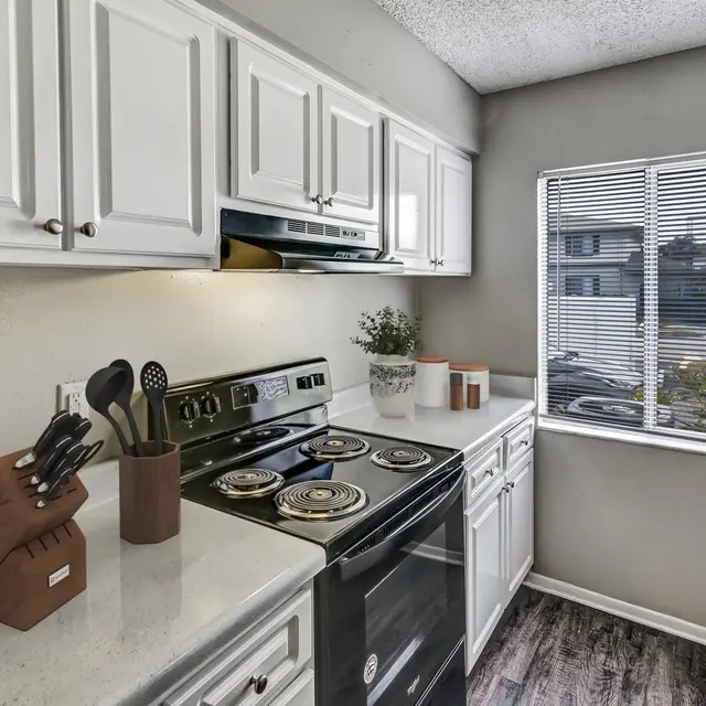 A modern kitchen with white cabinets, a black stove, and a window providing natural light. Kitchen utensils are neatly arranged in a holder and there are potted plants on the counter.