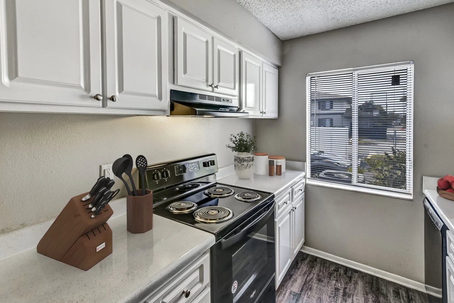 A modern kitchen with white cabinets, a black stove, and a window providing natural light. Kitchen utensils are neatly arranged in a holder and there are potted plants on the counter.
