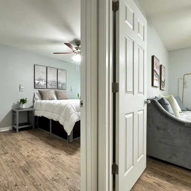 A view from a doorway into a modern bedroom, featuring two areas. On the left, a queen-sized bed with white bedding and framed art on the wall. On the right, a gray couch with decorative throw pillows and a floor lamp.