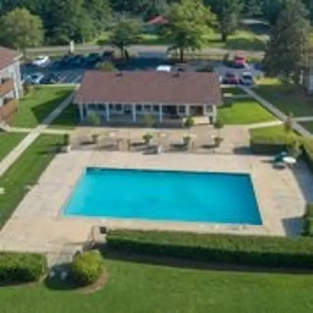 Aerial view of a community pool area surrounded by manicured grass and shrubs, with lounge chairs near the pool and a clubhouse in the background.