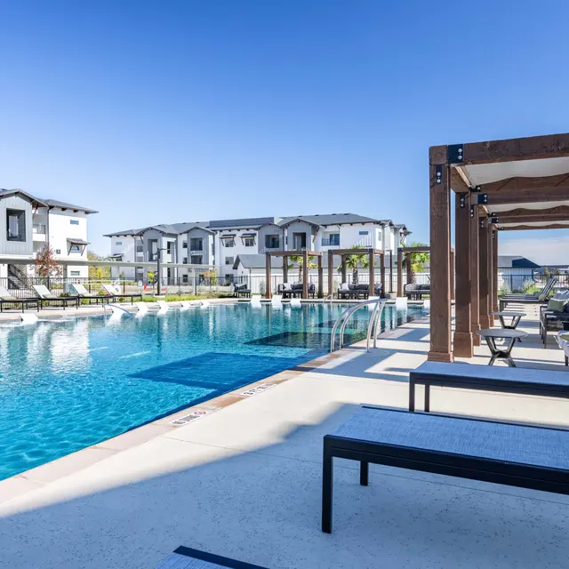 A luxurious outdoor swimming pool area at an apartment complex, featuring sun loungers and shaded cabanas, surrounded by modern buildings under a clear blue sky.