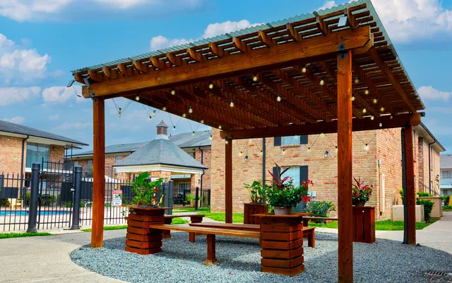 A wooden pavilion with a slatted roof, surrounded by small plants and gravel. It's located in a residential area with brick buildings and a fence in the background.