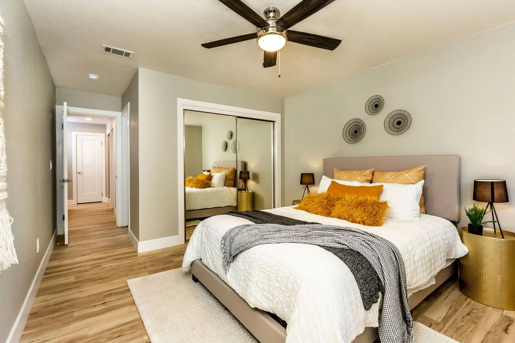 A modern bedroom featuring a bed with decorative pillows, a mirror, and a ceiling fan. There are warm wood floors and a path leading to a hallway with white doors.