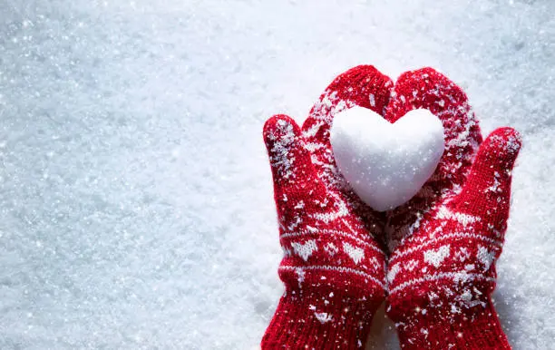 A pair of red mittens holding a white snow heart on a snowy surface.