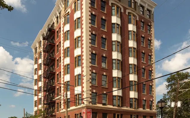 Historic Building Corner View A historic brick building with multiple stories, featuring large windows, fire escapes, and decorative cornices, situated at the corner of a street with cobblestones and parked cars.