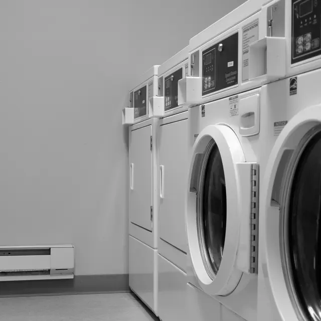 A laundry room featuring several white washing machines and dryers arranged along the wall, with a heating unit visible below. The image is in black and white, emphasizing the machines' clean lines and functionality.