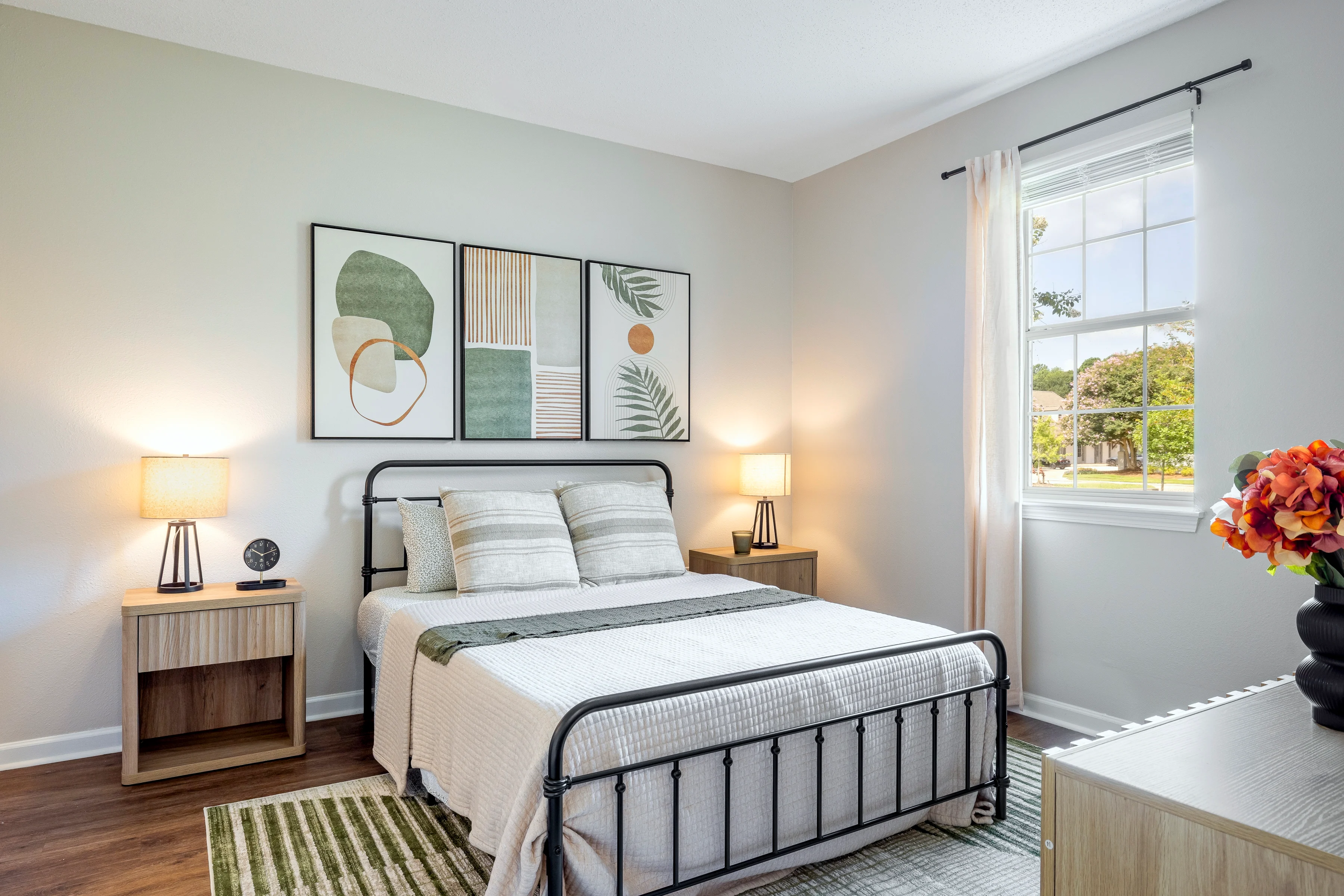 Modern Bedroom Interior A stylish bedroom featuring a metal bed frame with a white and green bedding set, two nightstands with lamps, and decorative wall art. A window allows natural light in, highlighting the minimalist decor.
