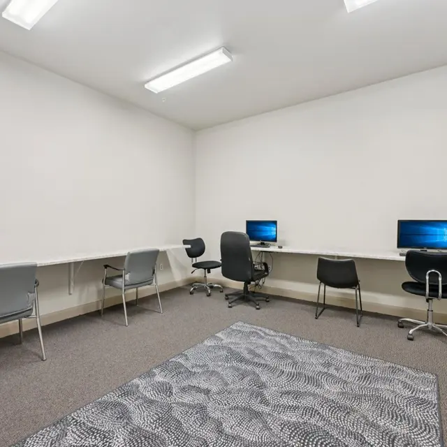 Modern Office Space A clean, minimalistic office space with two computers on desks, and several chairs arranged around. The room has a light grey carpet and plain white walls, illuminated by fluorescent ceiling lights.