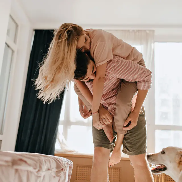 A couple playfully engaging together in a bright living room, with a dog nearby. The woman is sitting on the man's back, while he supports her by holding her legs, both smiling joyfully. Sunlight streams in through large windows, illuminating the scene.