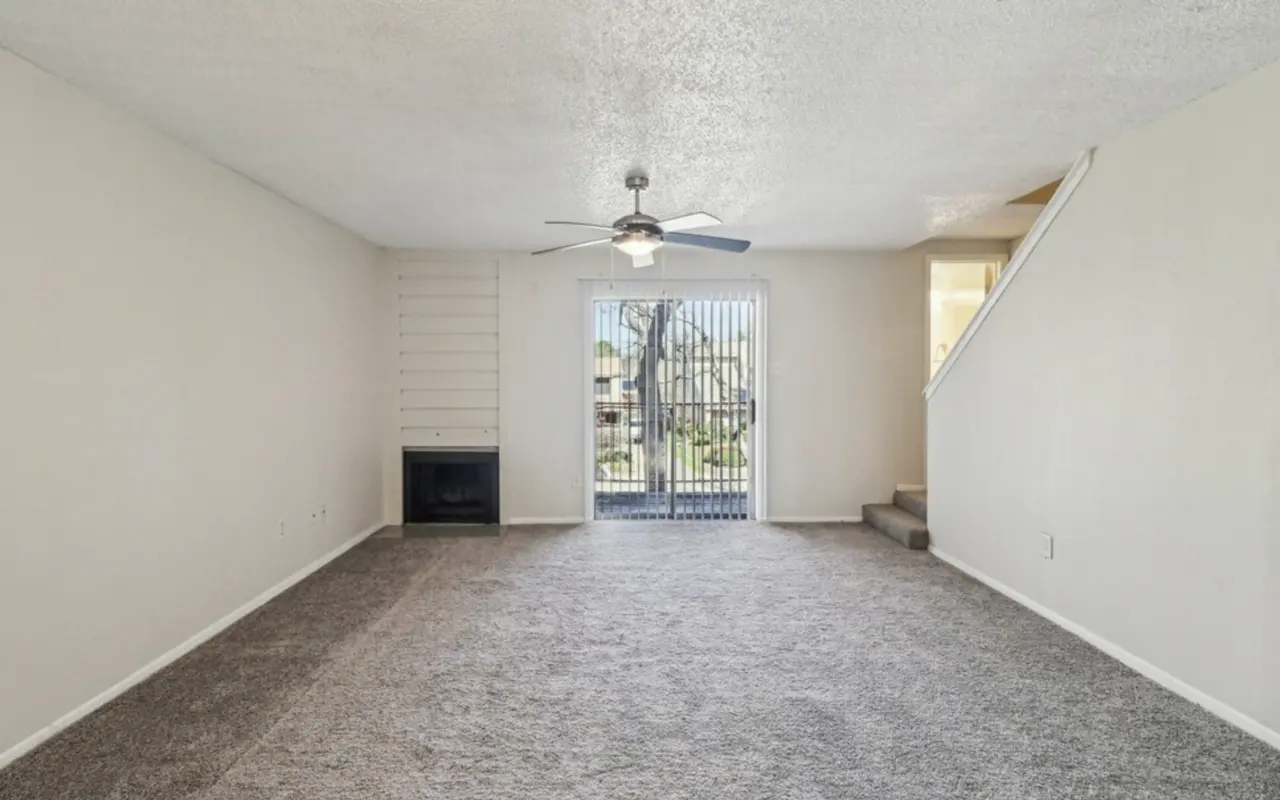 A spacious living room with light-colored walls and carpet, featuring a ceiling fan and a large window with metal bars letting in natural light. A staircase is visible in the background.