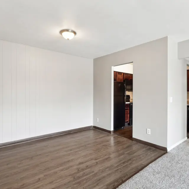 A spacious empty living room with hardwood floors, light gray walls, and a ceiling light fixture. The entrance to a kitchen is visible in the background, along with another room leading off to the right.